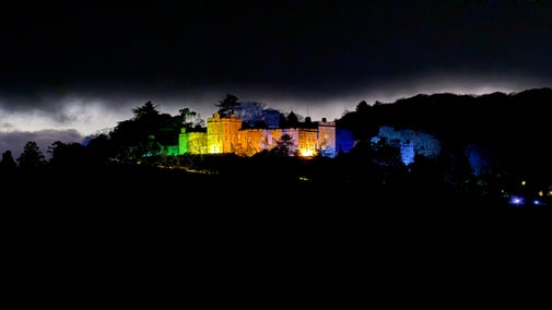 Dunster Castle illuminated at night with vibrant colored lights, including shades of yellow, green, and blue, highlighting the historic building against a dark sky. Silhouetted trees surround the castle, and a dramatic glow from distant clouds adds atmosphere to the scene.